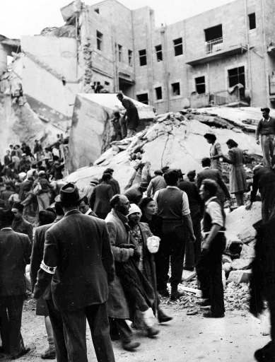 An injured Jewish couple walk past rescuers searching for wounded and dead people in the wreckage of shops on Ben Yehuda Street, Jerusalem, Feb. 22, 1948, after a bomb exploded. The bomb killed 52 Jews and wounded 100 more and levelled buildings on both sides of the street. 