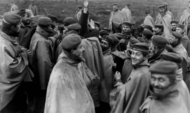 Cheerful German prisoners in forage caps and waterproof capes surround a member of a visiting News Agency in an effort to cadge cigarettes. Western Front, c1918. 