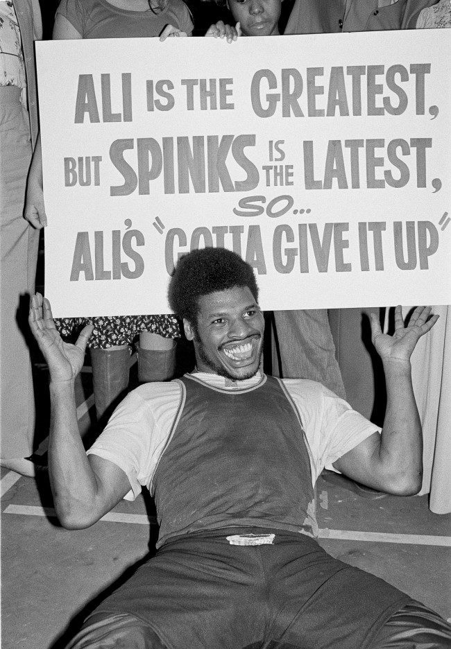 Heavyweight challenger Leon Spinks clowns for the photographers in Las Vegas, Feb. 10, 1978 where he is in training for his title bout against Muhammad Ali, Feb. 15. The sign was provided by some Spinks promoters. (