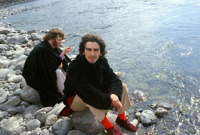 Beatles George Harrison and John Lennon, background, sit on rocks by a river in Rishikesh, India, in 1968. They are studying transcendental meditation with their yogi. (AP Photo) Date: 01/01/1968 