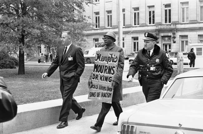 A plainclothes detective and a uniformed Shelby County Deputy sheriff usher away from the Shelby County Jail a picket who attempted to walk the sidewalk around the courthouse where James Earl Ray was scheduled to go on trial in Memphis, Tennessee on Nov. 12, 1968. Ray is accused of the murder of Dr. Martin Luther King.
