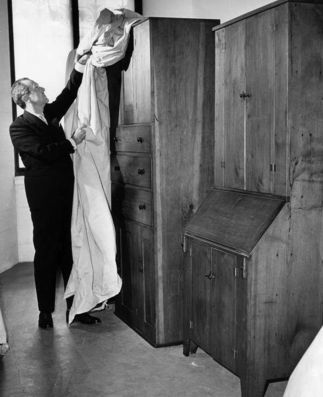 Henry McIlhenny, curator of decorative arts at the Philadelphia Museum of Art in Philadelphia, March 29, 1962, uncovers chest of drawers and slant-top desk made by members of the early-American religious sect known as the Shakers. The museum is preparing a month-long exhibit starting April 19 of what they believe will be the largest collection of Shaker crafts over assembled. The Shaker materials will be exhibited in six special galleries now under construction duplicating the plain residences of the sect. 