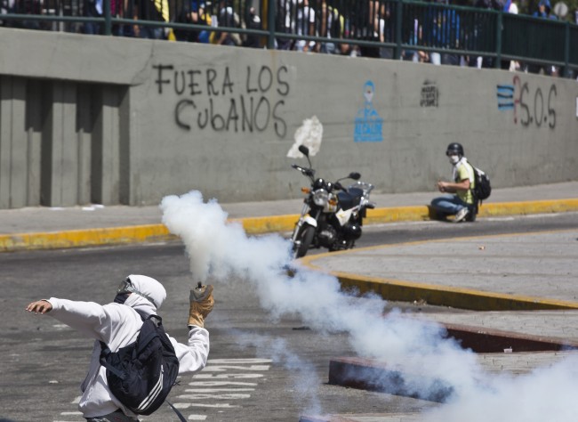 A masked protester returns a tear gas canister fired by Bolivarian National Police, in Caracas, Venezuela, Thursday, March 20, 2014. Thursday dawned with two more opposition politicians, San Cristobal Mayor Daniel Ceballos and San Diego Mayor Enzo Scarano, behind bars. Police used tear gas and water cannons to disperse a student-called protest of several thousand people in Caracas, some of those demonstrating against the arrests of the mayors.