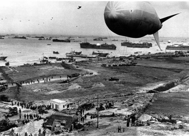 Allied materials of war, including amphibious "ducks" and other armored vehicles, fills an invasion beach on the Normandy coast of France, protected by barrage balloons during World War II. The balloons also hover over ships anchored off shore waiting their turn to unload the reenforcement supplies June 18th, 1944