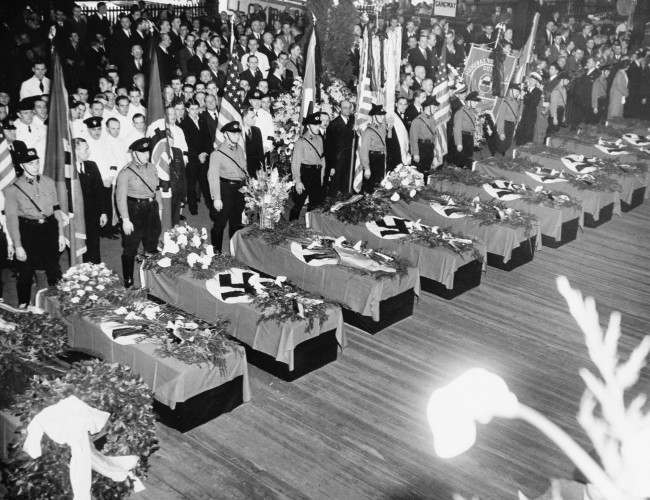 Funeral services for the 28 Germans who lost their lives in the Hindenburg disaster May 6 are held on the Hamburg-American pier in New York City. The swastika-draped caskets will be placed on board the Hamburg for their return to Europe. About 10,000 members of German organizations line the pier. 
