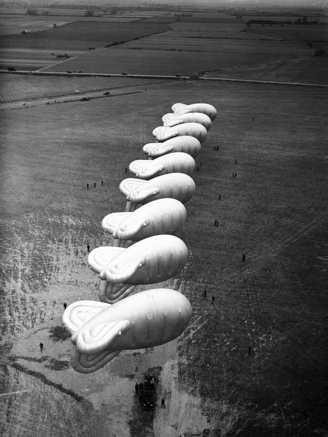 General view of balloons of the new balloon barrage groups of the Royal Air Force lined up in the air in Cardington, Bedfordshire, England on May 9, 1938. The balloons, attached to a great length of cable, ascend to an amazing height. The earth end of the cable is operated by a winch which can be mounted on a lorry to give the balloon mobility. Balloons, unused for air defence purposes since the war, will be used, if necessary for the protection of London from air attack. A number of these balloons will ascend from here on May 28, when Lord Luke, unseen, officially opens the empire air day. 