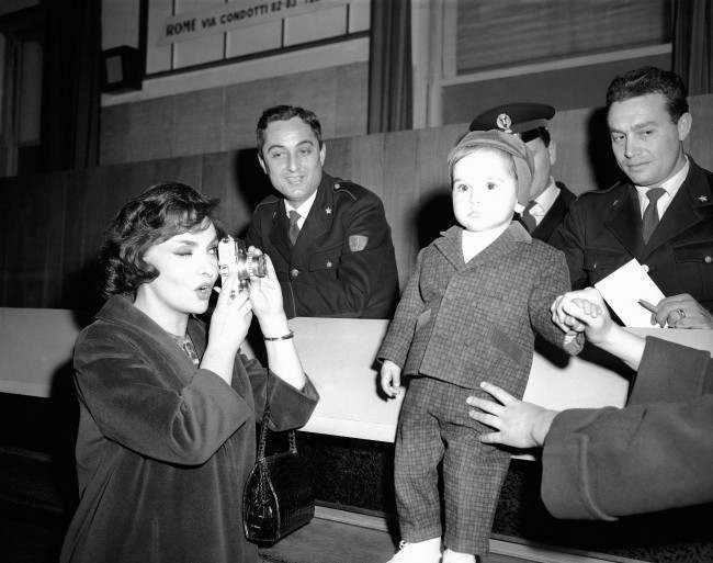 Like any other doting mother, actress Gina Lollobrigida takes pictures of her 19-month-old son, Milko Jr., at Rome's Ciampino Airport, March 13, 1959 as they awaited arrival of Gina's husband, Dr. Milko Skofic. Airport police were interested spectators. 