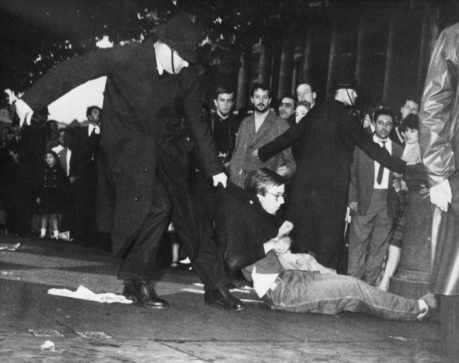 A young man in beatnik-style clothes is dragged along by a policeman