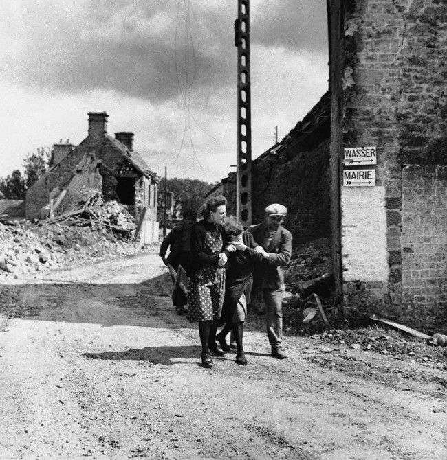 Two friends assist a weeping woman (center) in the Normandy village of Ste. Marcouf on June 19, 1944, after her husband was killed when the Germans shelled the place. The manÂs body is inside the store at right, upon which the Germans placed two signs: Wasser (German for water) and Mairie (French for town hall). (AP Photo)