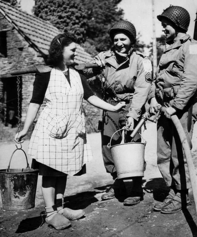 Two American soldiers, Staff Sgt. Bernard Dargols, New York, and Tech., 5/c William L. Stanley, Houston, Texas, right, help a French farm girl fill her water pails from an army water purification unit during a quiet moment somewhere in the Normandy beachhead area of France on June 30, 1944. (AP Photo)