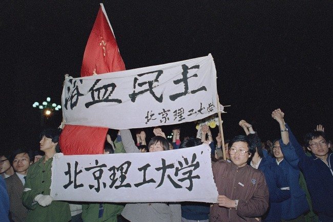 Students from Beijing University of Engineering carry a banner, top, reading 'Democracy, Bathed in Blood,' as they march into Beijing's Tiananmen Square to join tens of thousands of other students early on Saturday, April 22, 1989. The students plan to spend the night in the square and wait for the funeral of Hu Yaobang later in the day. (AP Photo/Mark Avery)