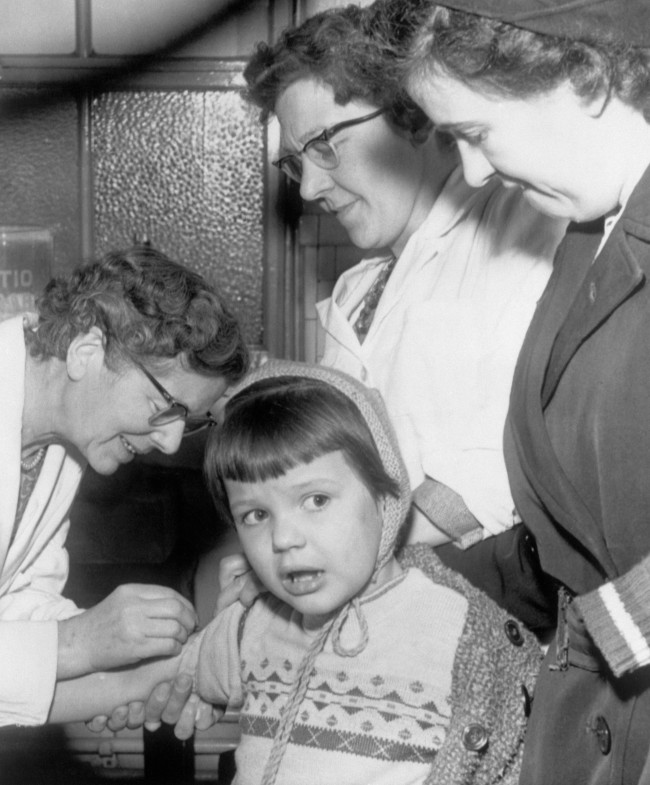A mother looks on as her child is vaccinated in a Bradford clinic as a precaution against smallpox, after an outbreak which claimed the lives of four people. Date: 14/01/1962 