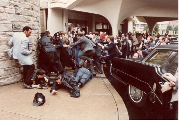 Police and Secret Service agents diving to protect American President Ronald Reagan amid a panicked crowd during an assassination attempt by John Hinckley Junior outside the Washington Hilton Hotel, Washington, DC.