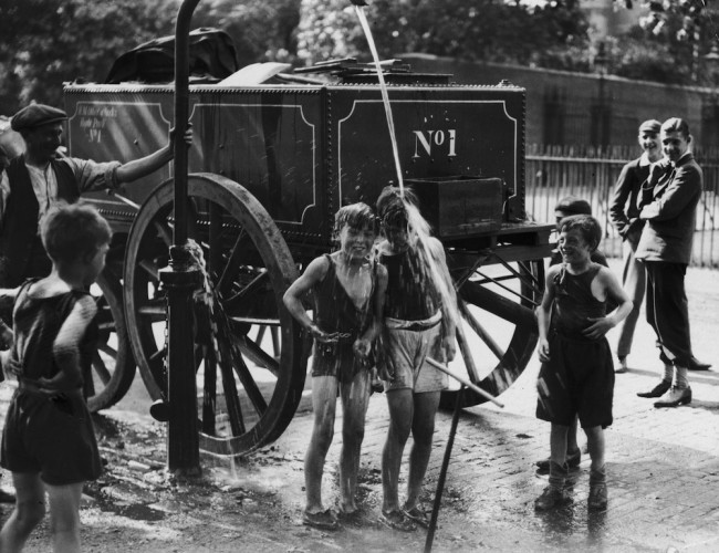 August 1930:  A water cart man turns the water main on a group of boys to help them cool off in a street in Westminster, London during a heatwave.  (Photo by Fox Photos/Getty Images)